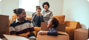 Young family of four smile and laugh together in a living room full of moving boxes.
